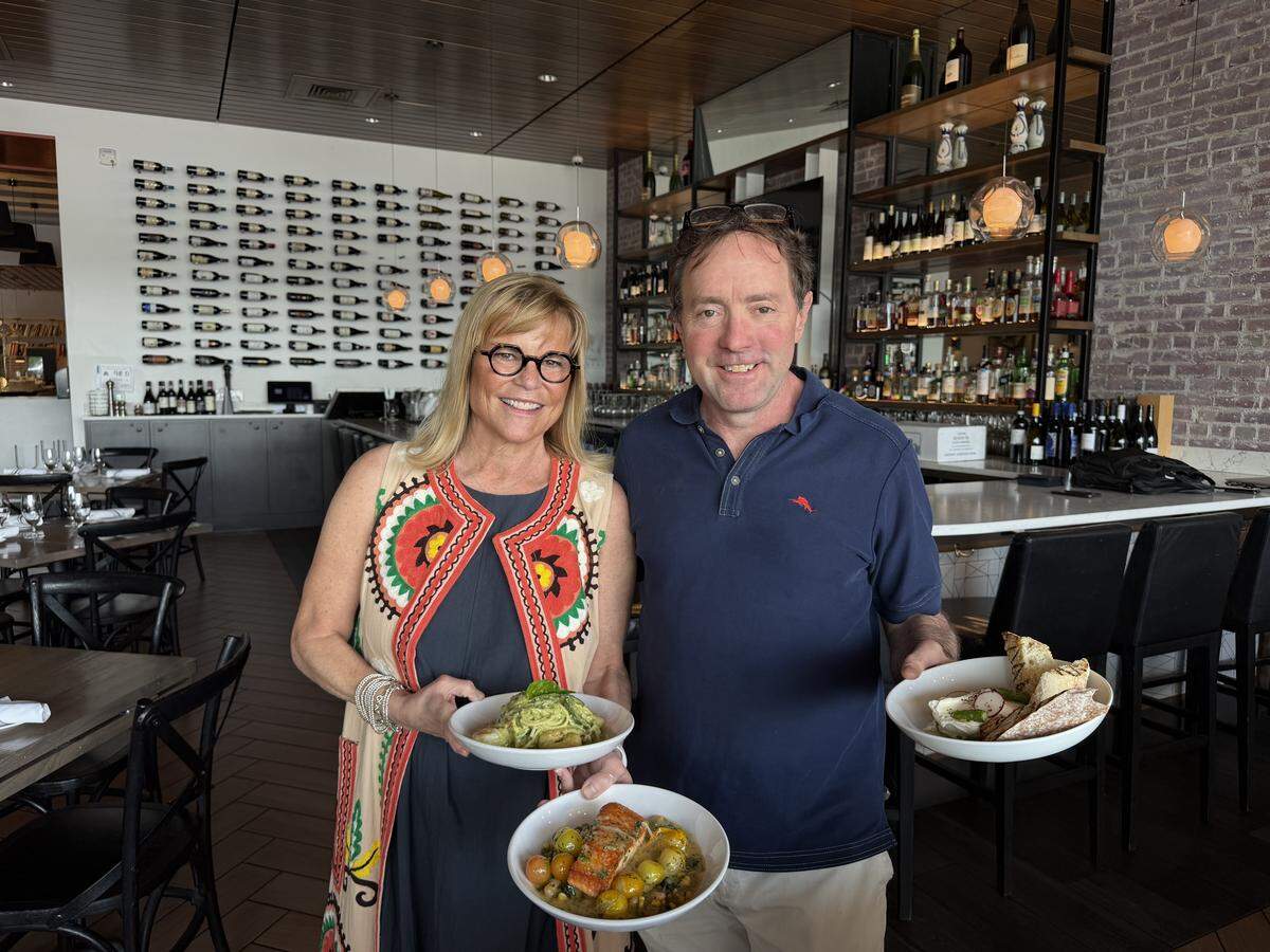 Two restaurateurs stand together inside their restaurant, each holding white bowls of prepared food. The one on the left holds a pasta dish and a salmon fillet with cherry tomatoes, while the other holds a plate with fresh burrata and grilled bread. They are positioned in front of a modern bar and an extensive wine rack.