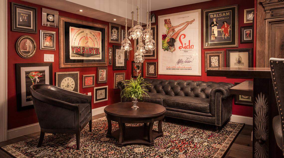 A luxurious, dimly-lit library or lounge with red walls completely covered in an eclectic gallery of framed vintage art and clocks. The room features a dark brown leather Chesterfield sofa, a matching club chair, and a round wooden coffee table. Multiple ornate glass chandeliers hang above the center of the room. The floor is covered by a patterned Oriental-style rug.