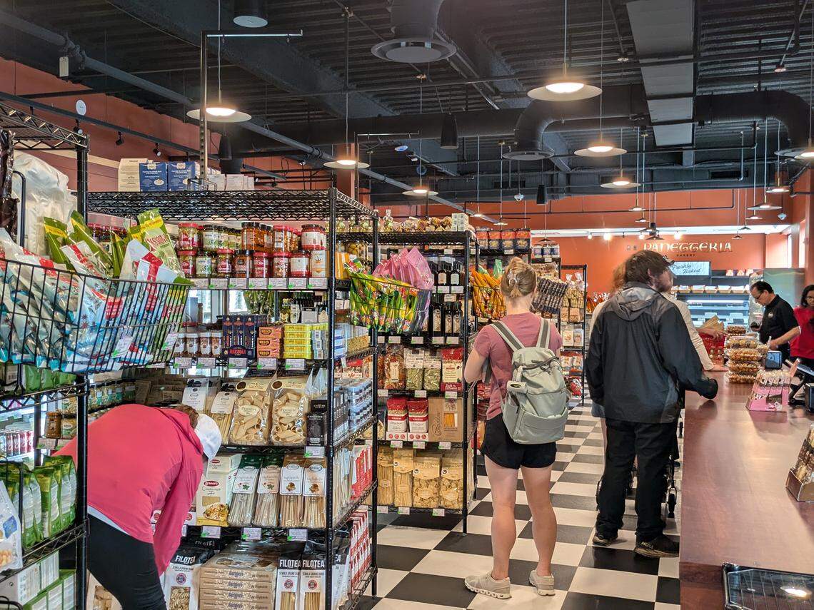 An interior view of the market aisles with a black and white checkered floor. Customers browse tall black wire shelves filled with imported Italian dry goods, including various pasta shapes, sauces, and oils.
