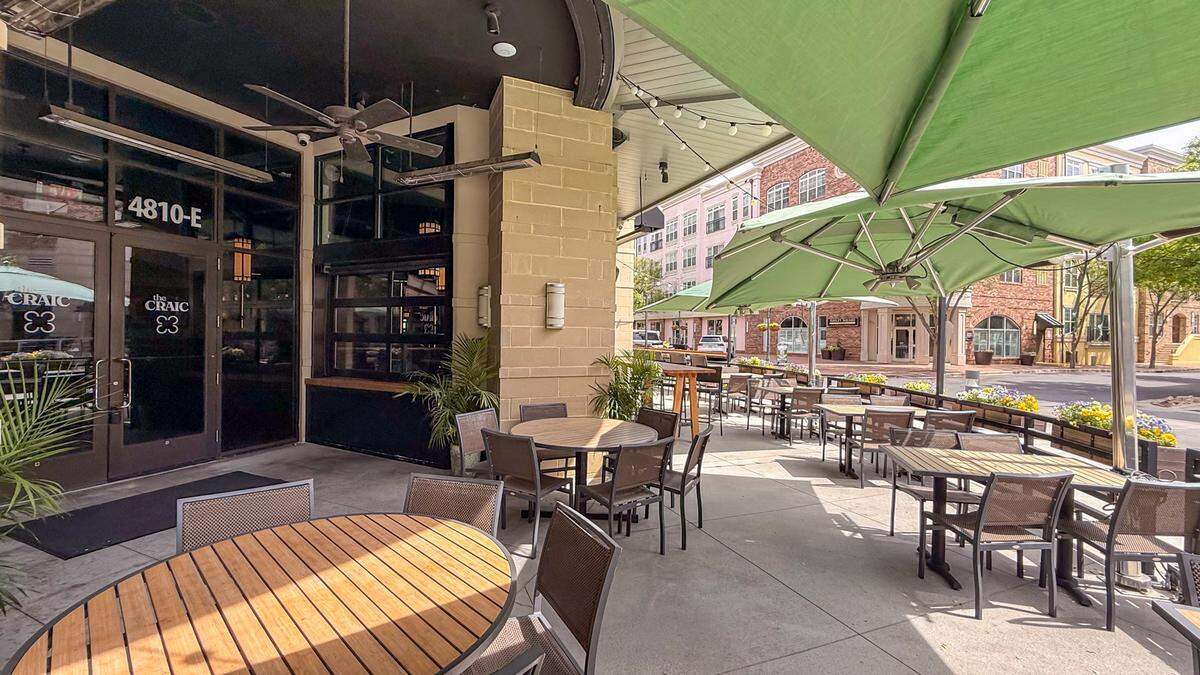A view of the pub’s exterior entrance and the full length of the patio. The glass entrance doors are labeled with “4810-E” and the pub’s name, “the CRAIC,” accompanied by a three-leaf clover logo. The patio extends along the building, featuring a mix of round and rectangular tables under green umbrellas. Overhead, string lights and industrial heaters are mounted to the underside of the building’s overhang, suggesting a space designed for year-round use.