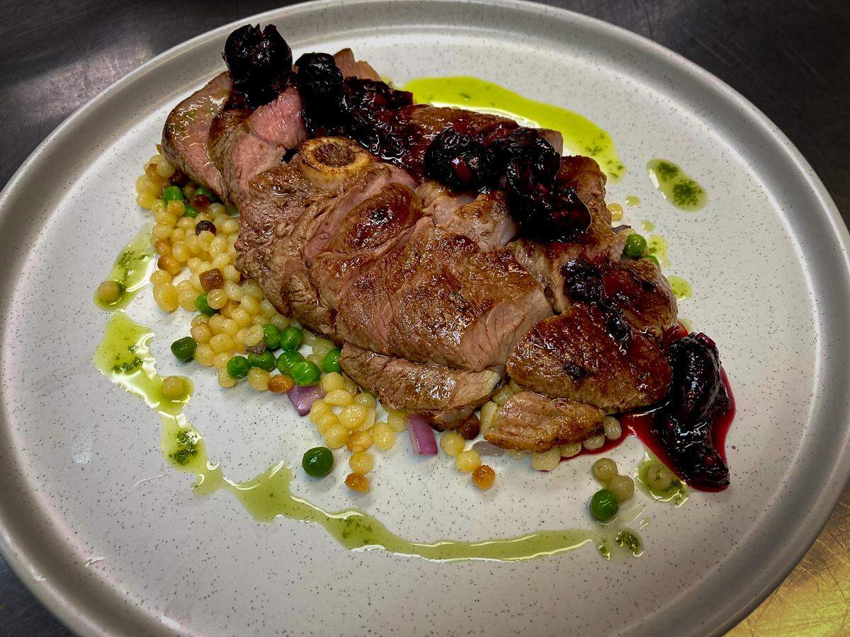 On a modern stoneware plate, a serving of roasted lamb is artfully arranged. The sliced, bone-in lamb has a seared exterior and pink center, and is garnished with a dark cherry compote. It is served over a bed of pearl couscous with peas and red onion, and drizzled with a green herb oil.
