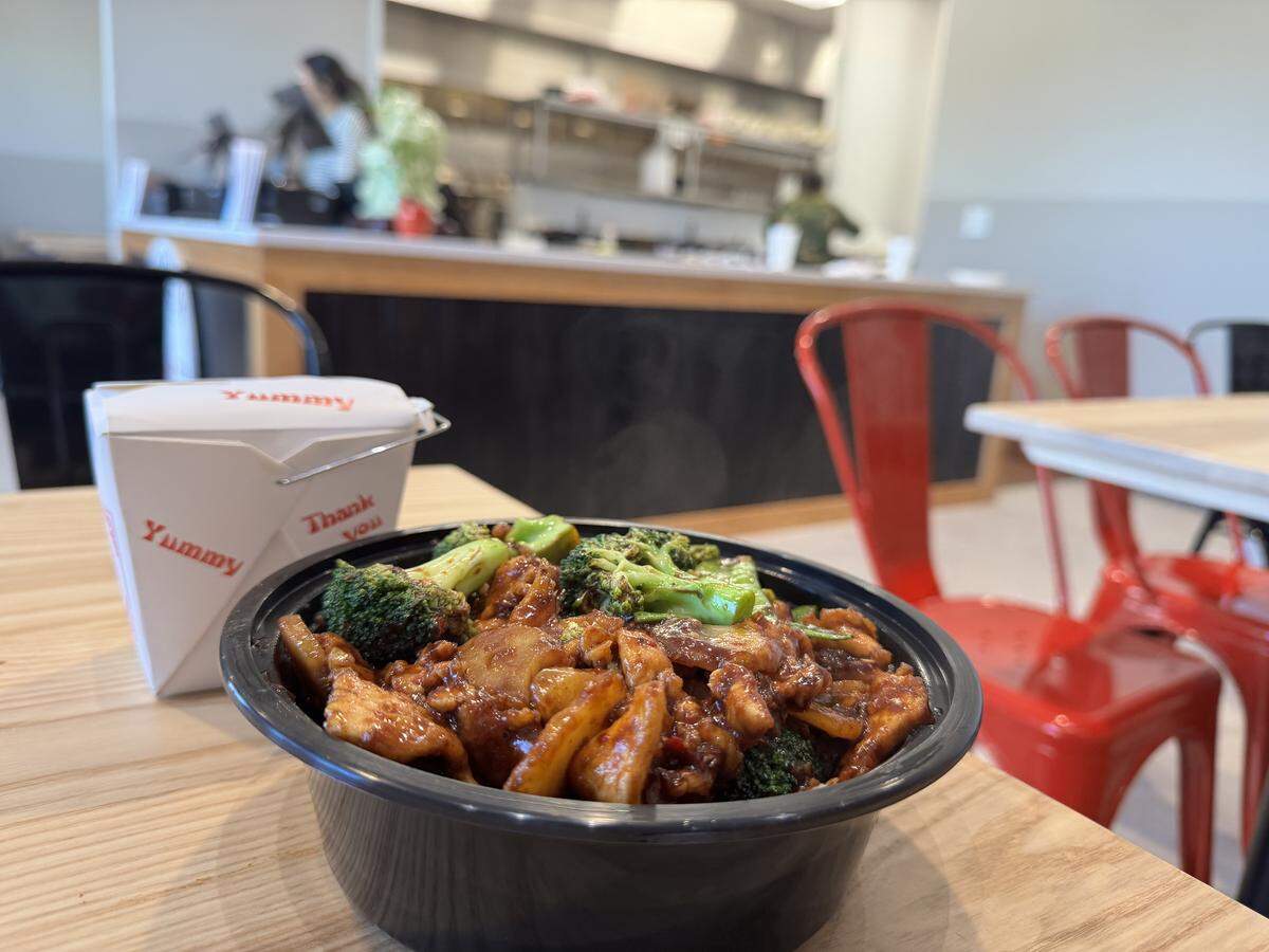 A dish of Sa Cha Chicken from Taipei Express is presented in a black bowl on a light wood table. The stir-fry contains sliced chicken and bright green broccoli in a savory sauce. A classic white takeout box for rice sits next to the bowl, with the casual, modern restaurant blurred in the background.