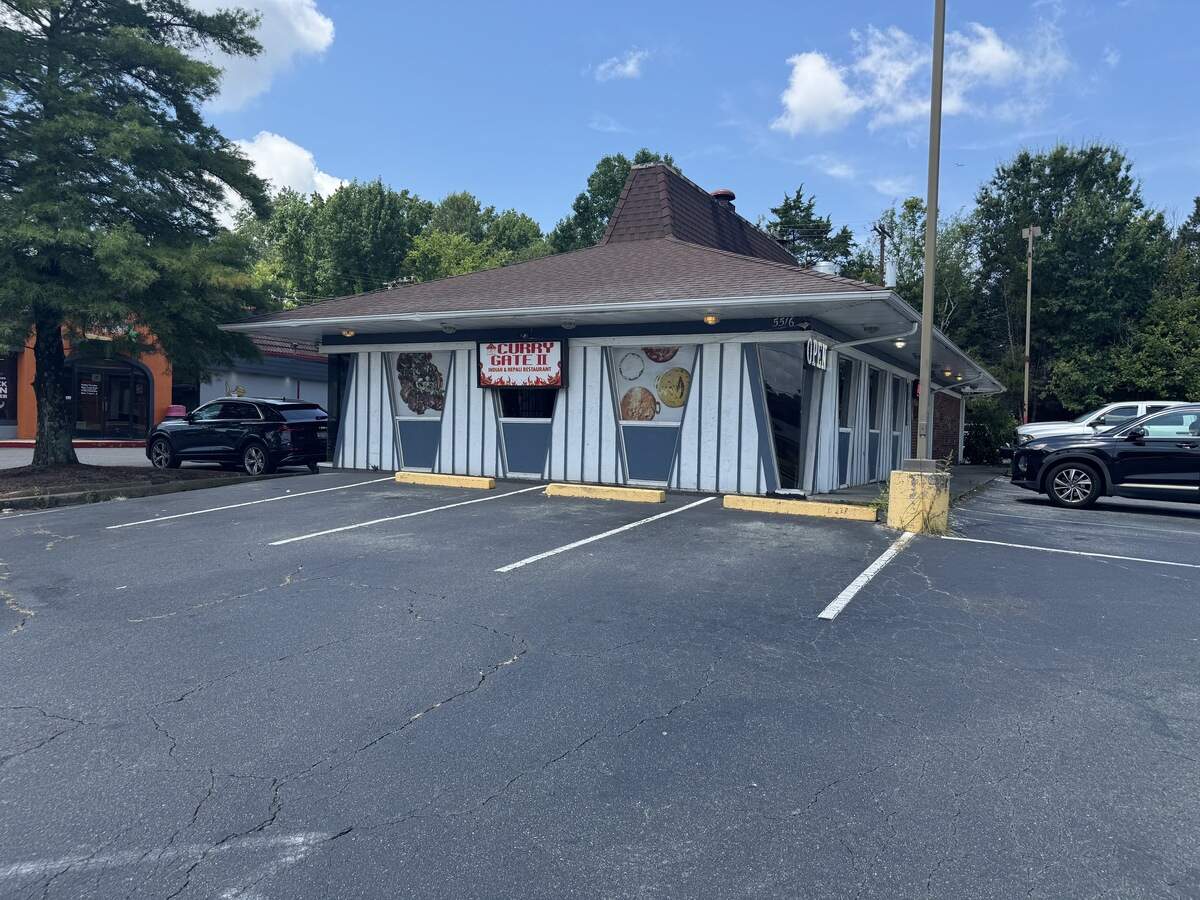 A wide exterior shot of an Indian and Nepali restaurant named “Curry Gate II” on a bright, partly cloudy day. The restaurant is housed in a unique, retro building featuring a steep, brown-shingled mansard roof and angled white-and-gray paneled exterior walls. A black SUV and a black truck are parked on either side of the empty asphalt parking lot in front of the building.