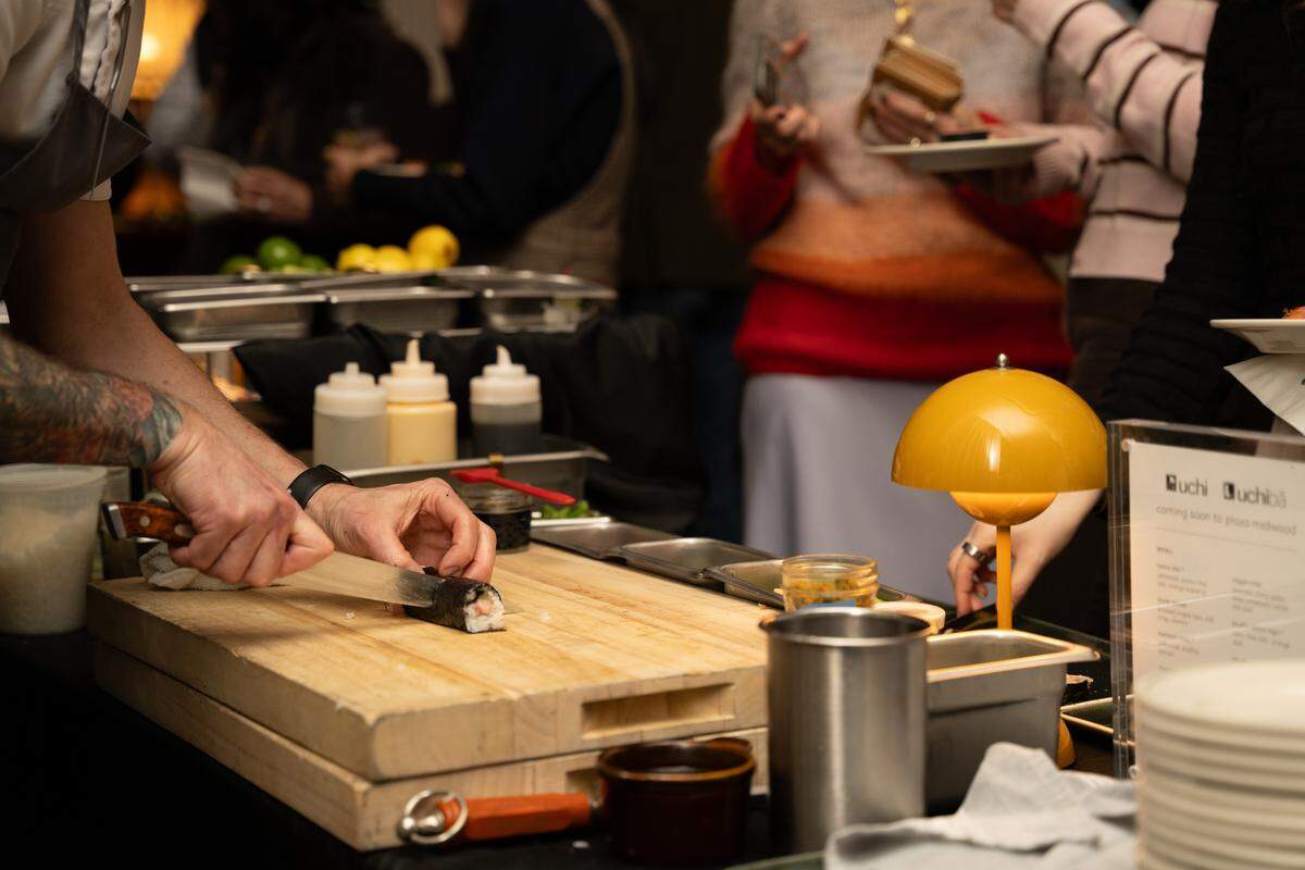 A close-up shot of a chef with tattooed arms using a professional knife to slice a sushi roll on a thick wooden cutting board. In the background, guests are blurred, and a small yellow dome lamp illuminates the workspace.