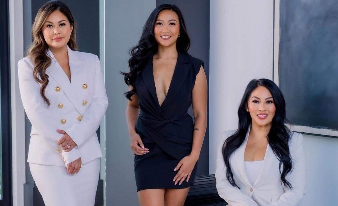 A portrait of three stylish women posing indoors. The woman on the left wears a white double-breasted blazer and a matching skirt with gold heels. The woman in the center wears a black, short-sleeved dress with a V-neck and black open-toed heels. The woman on the right sits on a bench, wearing a white blazer and matching pants with white pointed-toe heels. All three are smiling at the camera. The background is a modern room with dark gray walls and a large abstract painting.