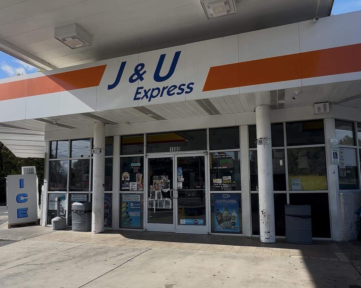 A straight-on, eye-level wide shot of the “J & U Express” gas station convenience store entrance. The white building, set under a large canopy with an orange stripe, features large glass windows and “J & U Express” printed in blue text. An “ICE” merchandiser is visible to the far left.