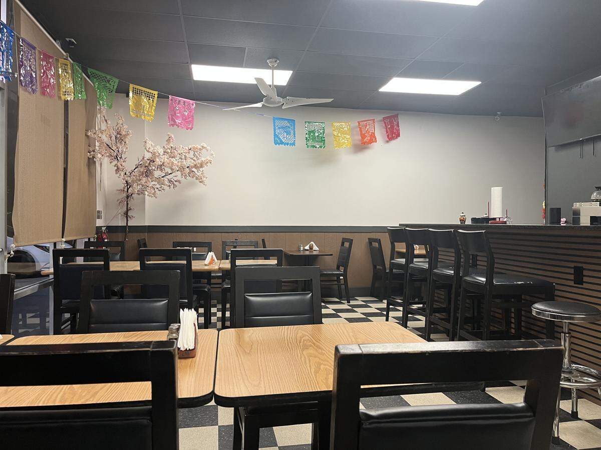 The interior of a casual restaurant, likely “Como en Mexico,” showing the dining area and a counter. The room has a black ceiling with bright square fluorescent lights and a white ceiling fan. The floor features a black-and-white checkerboard tile pattern. In the foreground, there are square wooden tables with black-framed chairs and black upholstered seats. Along the back wall, a bar-height counter with dark horizontal paneling is visible, fronted by black stools with backs. Colorful Mexican papel picado banners hang across the white wall, near a decorative, faux cherry blossom tree on the left.
