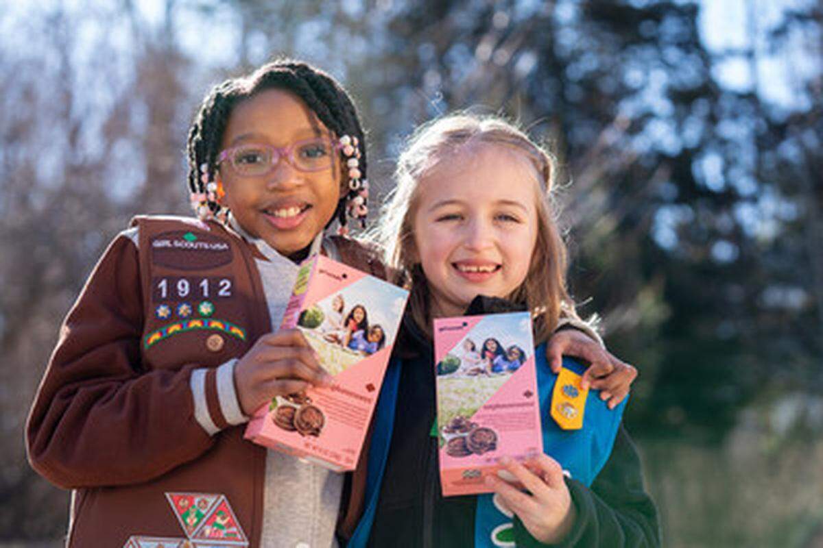 Two young Girl Scouts smile warmly at the camera while posing closely together outdoors. They each hold up a pink box of Exploremores cookies, proudly showcasing their participation in the cookie program.