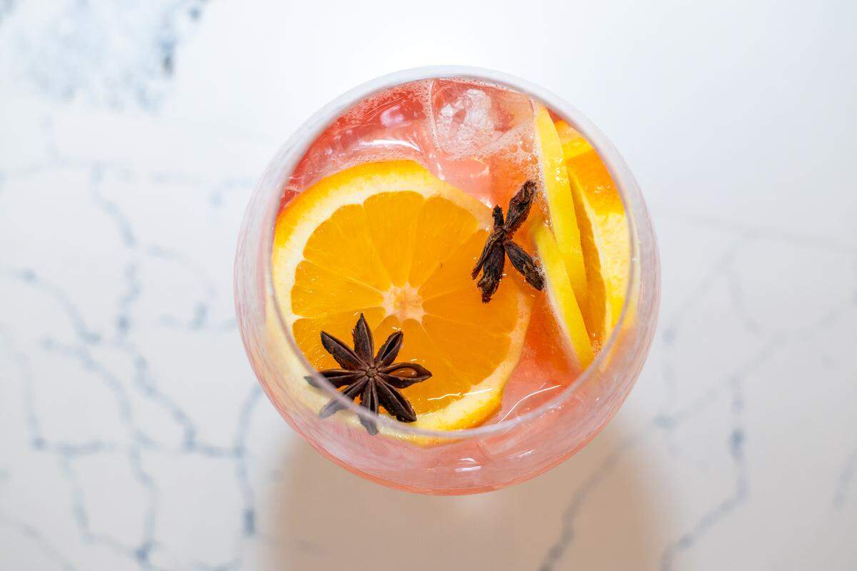 An overhead close-up shot of a round, glass of a pink or reddish cocktail. The drink contains ice and is garnished with a bright orange slice and two dark brown, star-shaped star anise pods. The glass is resting on a white surface with gray marble veining.