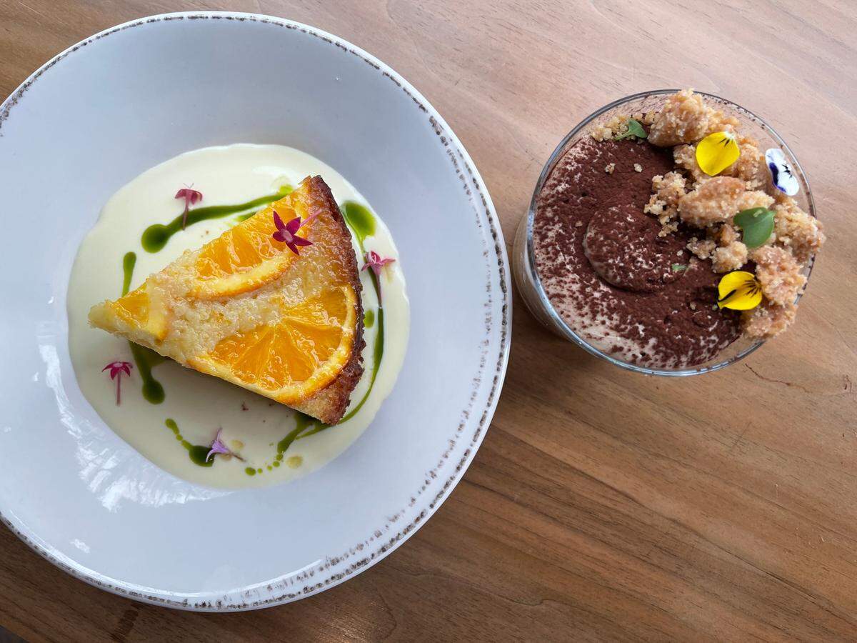A top-down view of two desserts on a light wooden table. On the left, a slice of orange olive oil cake, topped with thin orange slices and small pink edible flowers, rests on a pool of white cream sauce. Green sauce is drizzled around the cake, with small flower petals scattered on the plate. On the right, a glass of tiramisu is visible, dusted with cocoa powder and topped with crumbled cookies, a few green candied pieces, and bright yellow and purple edible flowers.