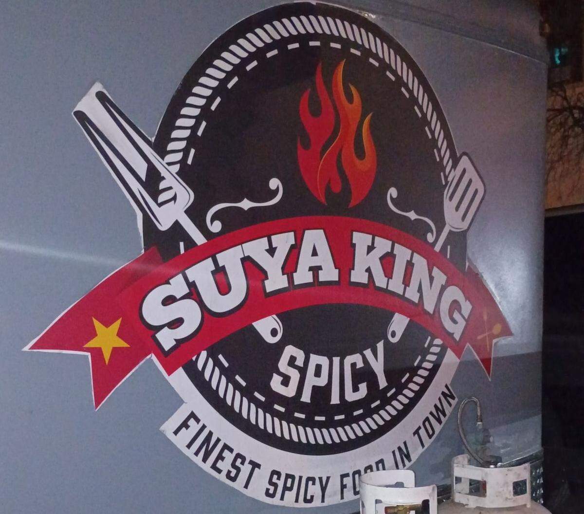 A close-up shot of a circular logo for a food business titled “SUYA KING”. The logo is displayed on the side of what appears to be a food truck or trailer. Key elements include the word “SUYA KING” written in large, bold, white letters across a curved red banner that features a yellow star on the left side. Below this, the word “SPICY” is printed in white, and a white border at the bottom contains the slogan “FINEST SPICY FOOD IN TOWN”.