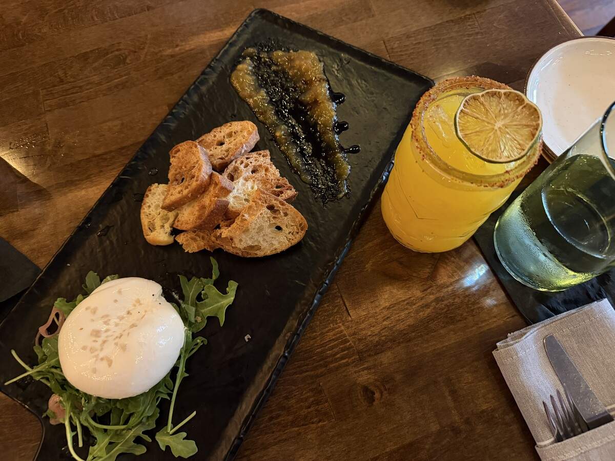 Burrata cheese on a bed of greens served with crostini, jam, and an orange cocktail on a wooden table.