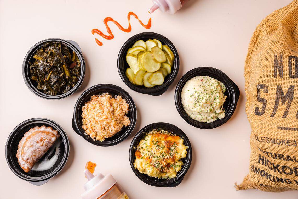 A flat lay photo showing five small black bowls filled with various barbecue side dishes, including collard greens, sliced pickles, potato salad, creamy rice, and baked mac and cheese, arranged around a fried hand pie. A burlap sack labeled “NOBLE SMOKE” is on the right.