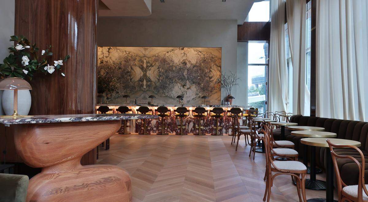 A sophisticated and spacious restaurant interior with modern design elements. The room features a light wooden herringbone floor. On the left, a large, dark wooden cylindrical pillar stands next to a uniquely shaped wooden table with a marble top, adorned with a lamp and white flowers. In the center, a long bar area is visible with multiple dark barstools lined up against a wall featuring an expansive, abstract art piece with natural tones. Beyond the bar, large windows are draped with tall, light-colored curtains, letting in natural light. On the right side of the room, several small round tables with dark bases and light chairs are arranged next to a long, dark cushioned banquette.