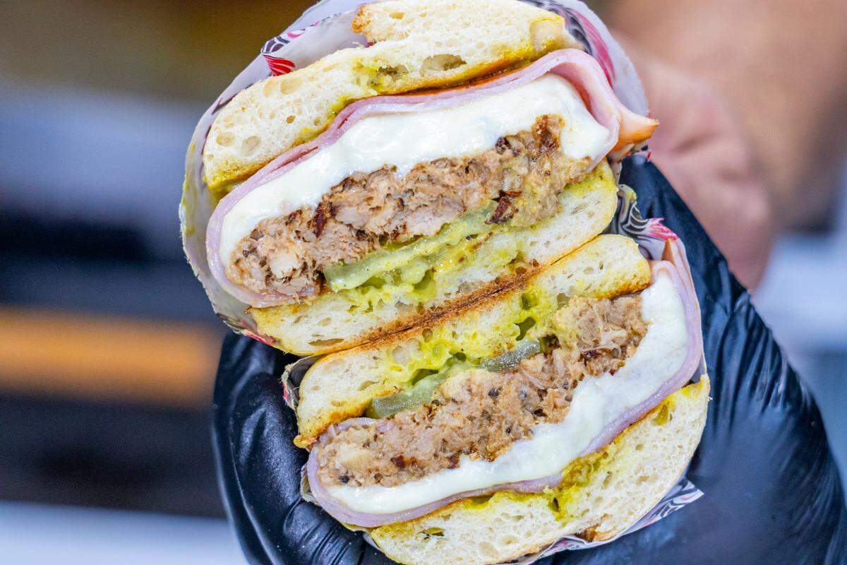 A close-up, overhead shot of black-gloved hands holding a sandwich, cut in half and stacked. The cross-section clearly displays thick, round patties of seasoned pork, layered with sliced ham, melted white cheese, pickles, and yellow mustard, all on a soft, round roll.