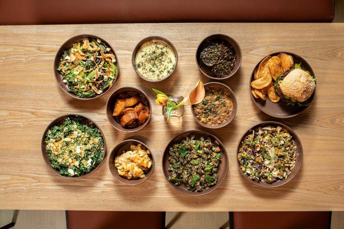 A top-down view shows a wooden table laden with various dishes in brown bowls. In the top row, from left to right, there’s a salad with greens and what looks like fried onions, a creamy white dish sprinkled with green herbs, a dark stew or grain dish, and a plate with a burger and potato chips. In the middle row, there’s a small glass vase with a single light orange flower and green leaves. Surrounding it are smaller bowls: one with roasted orange chunks (possibly sweet potatoes), and another with a bean or lentil stew. In the bottom row, from left to right, there’s a vibrant green salad with a yellow crumbly topping, a bowl with what appears to be macaroni and cheese or a similar pasta dish, a grain salad with green vegetables, and a slaw or salad with shredded vegetables. The table is light wood, and dark brown chairs are partially visible at the bottom of the frame.