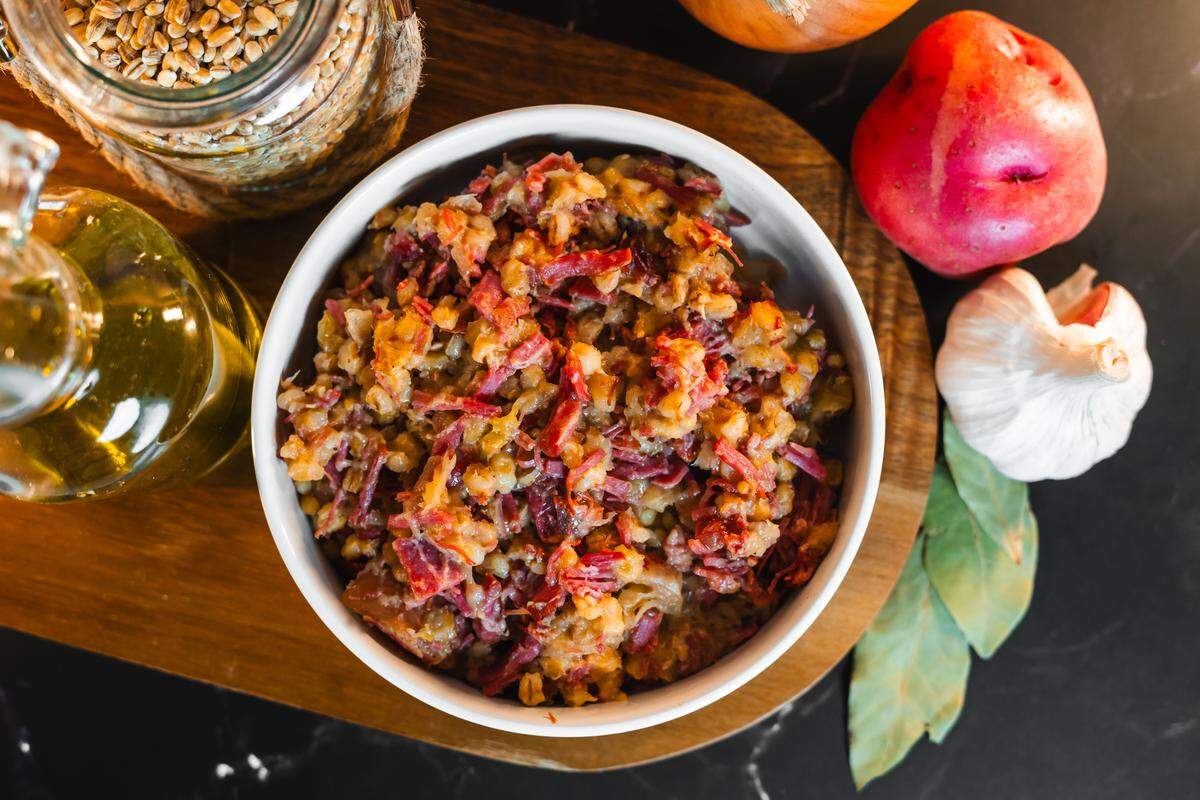 A top-down view shows a white ceramic bowl filled with a hearty corned beef hash mixed with shredded potatoes. The dish sits on a wooden serving board surrounded by raw ingredients, including a whole red potato, a head of garlic, loose bay leaves, and a glass jar of grains. A bottle of olive oil rests on the side, all set against a dark marbled countertop.