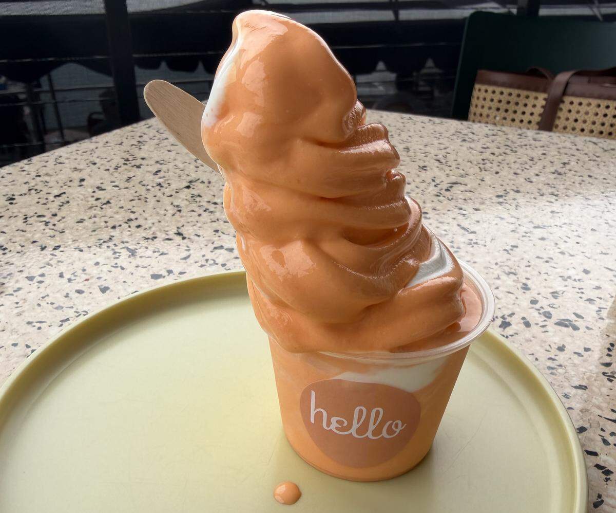 A tall swirl of orange and white swirled soft-serve ice cream in a clear plastic cup with a “hello” logo sticker. A wooden spoon is tucked into the side, and the cup sits on a pale green tray over a speckled stone tabletop.