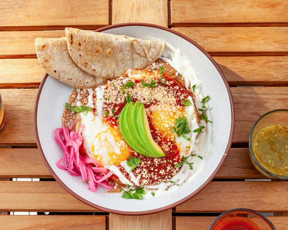 A top-down, close-up shot of a plate of Huevos Rancheros on a light-colored wooden slatted table. The dish is presented in a white speckled bowl with a brown rim.