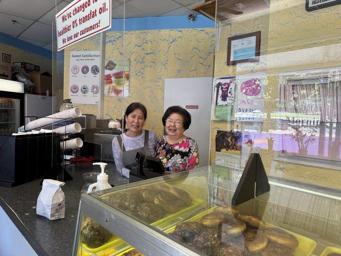 Posing for a photo from behind the counter of their shop, two smiling workers are seen. The one on the left wears an apron, and the one on the right wears a floral shirt.