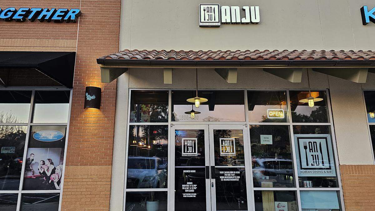 The exterior storefront of Anju, a Korean dining and bar. The restaurant is located in a modern strip mall with large glass windows and a door. A sign above the entrance displays the restaurant’s name and logo, which is also visible on the glass. A lit “OPEN” sign is visible inside.