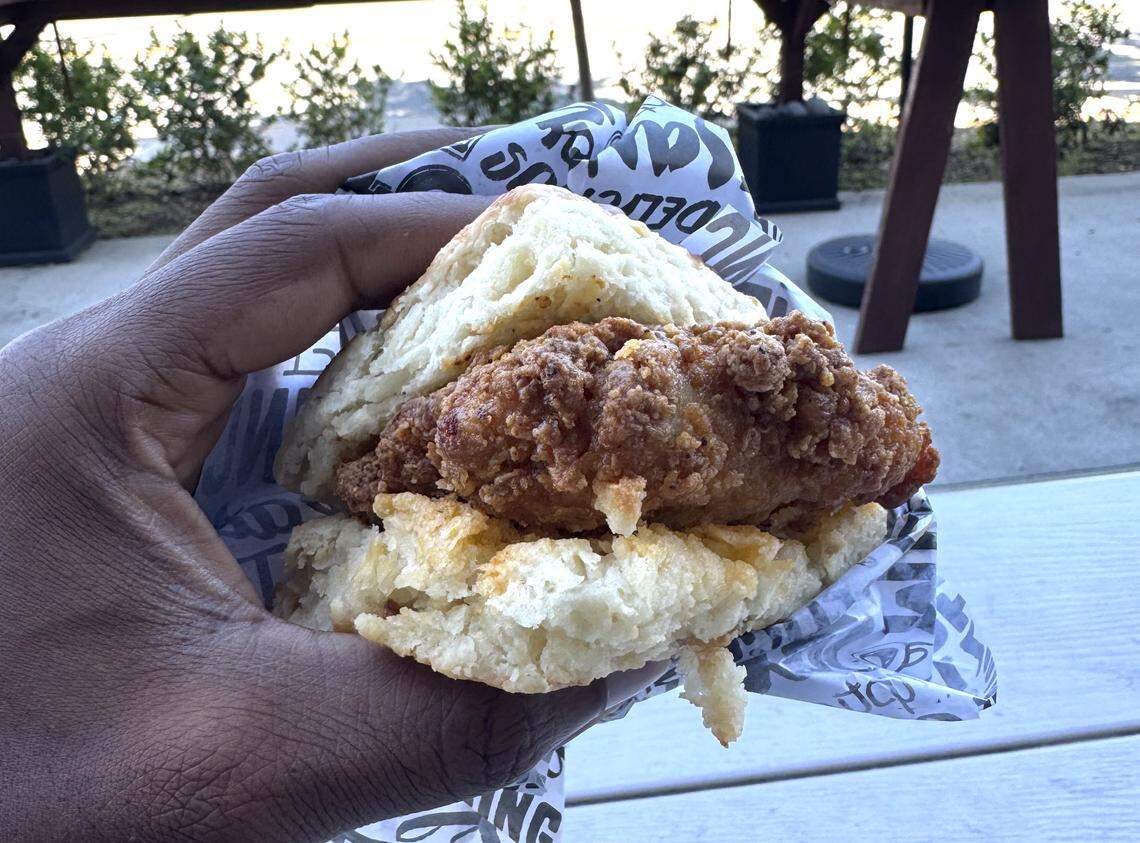 A close-up, first-person view of a hand holding a large fried chicken biscuit wrapped in patterned deli paper. The biscuit is fluffy and golden, and the chicken breast is thick with a craggy, crispy breading. The background shows a sunny outdoor patio with wooden tables and green trees.