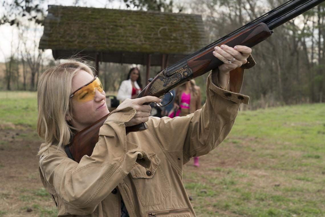 A blonde gun user in a tan jacket is aiming a shotgun with an ornate, engraved receiver, wearing yellow-tinted shooting glasses, cheek pressed against the stock. In the blurred background, another shooter in a white jacket and a third in a pink jacket are visible, standing near a covered wooden structure. The setting appears to be a grassy field with trees in the distance.