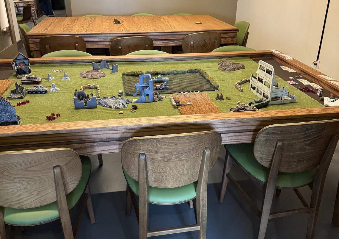 A close-up shot of a large, custom-built wooden wargaming table with a green playing surface representing a battlefield. The table is covered in miniature terrain, including tanks, infantry models, dice, and scenery like a large white ruined building and smaller concrete barriers, with wooden chairs pulled up around it.