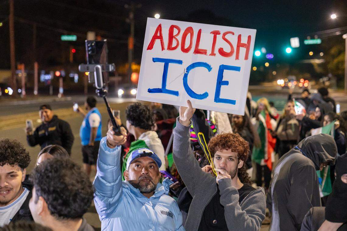 A group of approximately 20 protesters stands across a lane of a dark, wet street at night. They are prominently holding several large Mexican flags, which are the main focus of the image. One protester in the foreground holds a smaller flag with multiple red and white symbols. Arrow markings are painted on the asphalt, and car headlights are visible in the background.