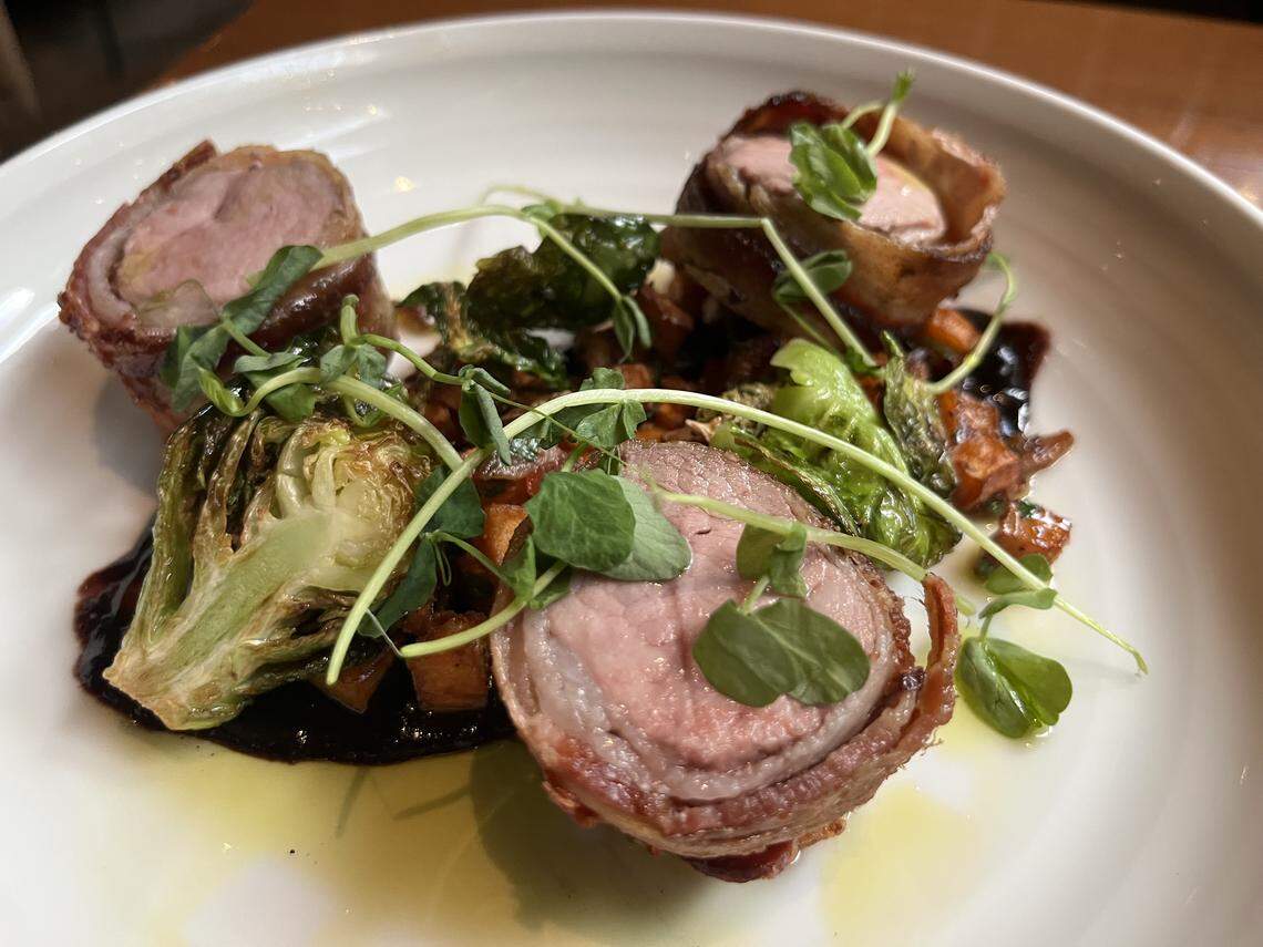 A close-up, high-angle shot of a plated entree. The dish features three pieces of pork tenderloin, each wrapped in bacon and cooked to a medium-rare pink. The pieces are arranged on a bed of roasted vegetables, which appear to be sweet potatoes and brussels sprouts. A dark, rich sauce is drizzled beneath the food, and the entire dish is garnished with fresh pea shoots. The meal is served on a wide, white plate.