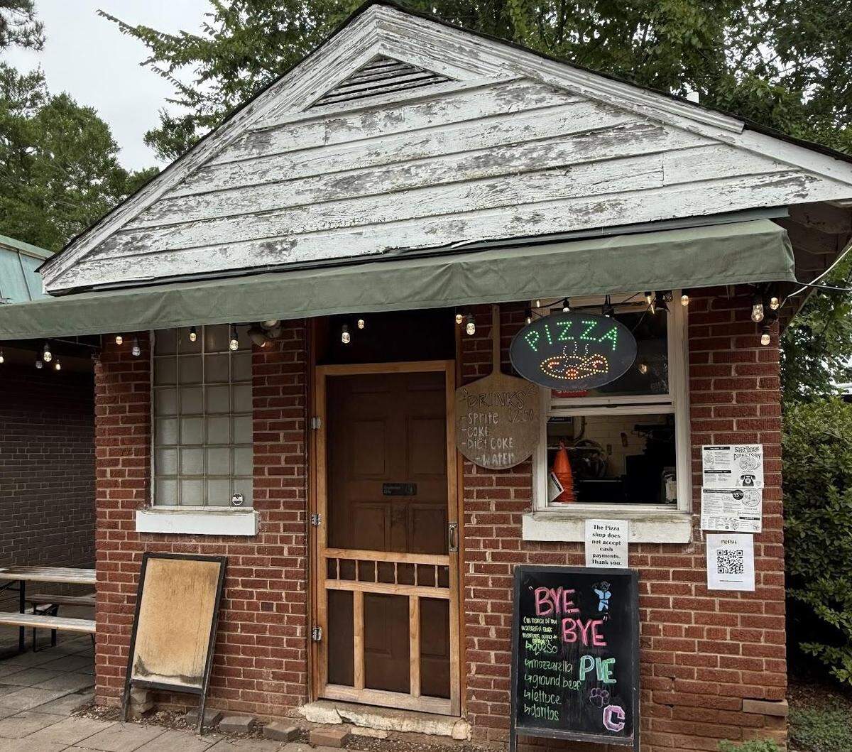 A detailed shot of a small brick building features a weathered gable with peeling white paint, a green awning, a screen door, and a service window. A neon pizza sign, a chalkboard menu, and other handwritten signs contribute to its quirky, local character.