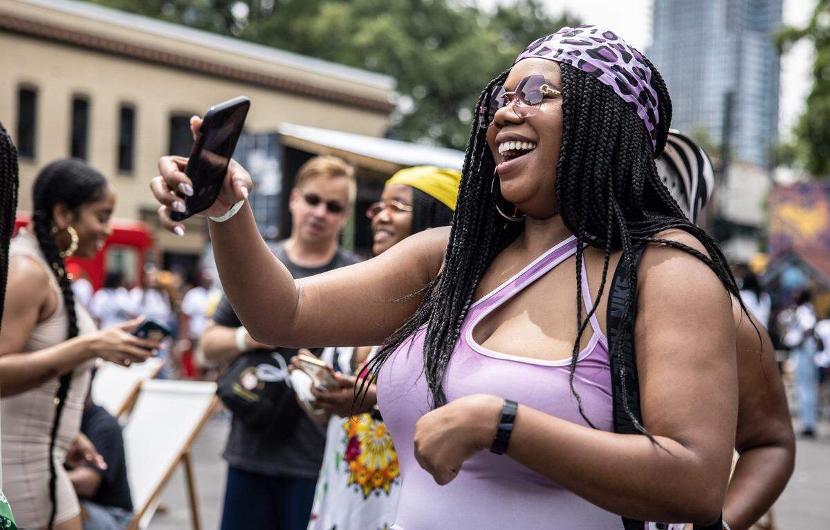 A Black person with glasses laughs joyfully while taking a selfie at a crowded outdoor festival while wearing a purple leopard-print headwrap over long, black box braids and a light purple bodysuit with a thin strap detail.