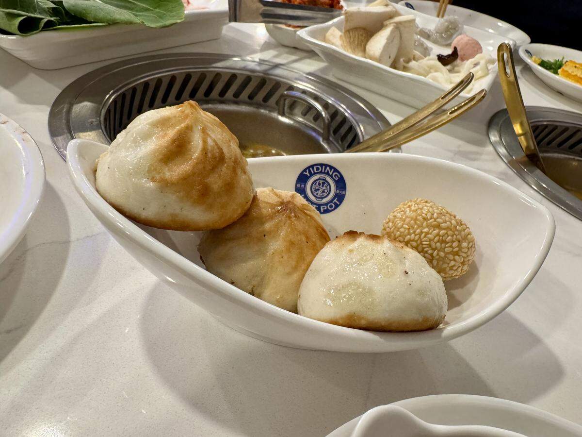 An appetizer from Yiding Hot Pot’s all-you-can-eat menu is shown in a curved white bowl. The bowl contains four small, pan-fried pork buns and one sesame ball. In the background, a hot pot burner and plates of raw ingredients for cooking are visible on the table.