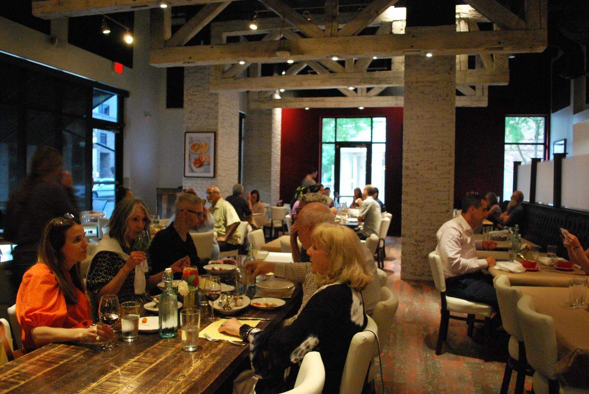 The interior of a busy restaurant filled with patrons dining at long wooden tables and smaller booths. The space features a rustic-industrial design with prominent white timber ceiling beams, exposed brick pillars, and warm, atmospheric lighting.