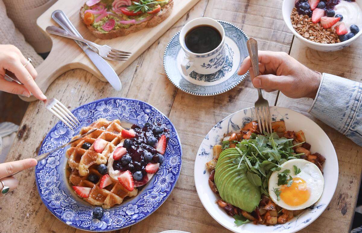 An overhead shot of a wooden table filled with brunch dishes, including a waffle topped with berries and cream, avocado toast, a breakfast bowl with a fried egg and sliced avocado, and a cup of black coffee. Two people are captured from above, using forks and knives to enjoy the colorful meal.