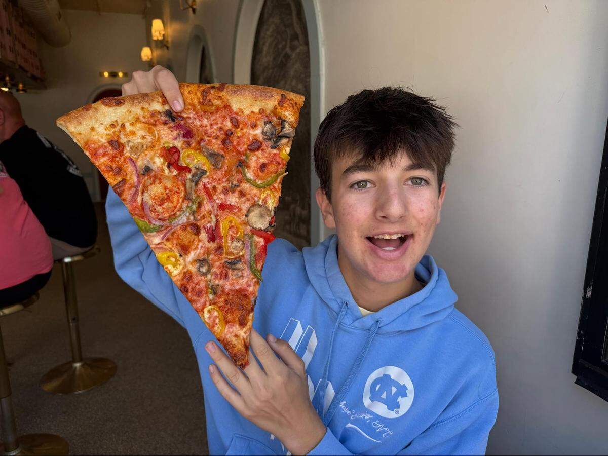 An eye-level, medium close-up shot of a happy teenager holding up a massive slice of New York-style pizza while wearing a light blue UNC Chapel Hill hooded sweatshirt and smiling widely. The oversized slice is loaded with cheese, red sauce, mushrooms, green peppers, red onions, and yellow banana peppers. The interior background of the pizzeria is softly blurred.