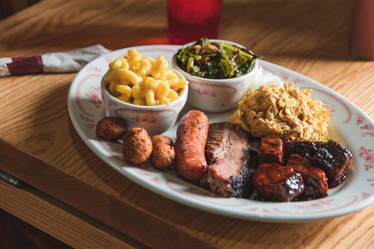 A hearty barbecue platter from Midwood Smokehouse is presented on a white oval plate with a subtle floral pattern. The plate holds a variety of meats and sides. Meats include a thick slice of brisket with a smoke ring, several pieces of dark, glazed burnt ends or pork belly, and a piece of sausage. Sides are served in small white bowls: a creamy macaroni and cheese and a bowl of green collard greens. A small pile of pulled pork and three hushpuppies are also on the plate. The meal is set on a light wooden table, with a blurred red glass visible in the background.