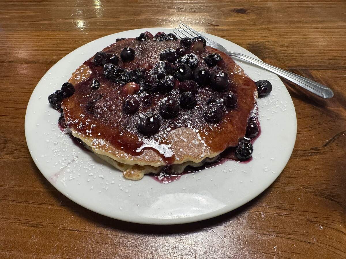 A stack of golden-brown pancakes served on a white plate, topped with blueberry syrup, whole blueberries, and a light dusting of powdered sugar, with a silver fork resting on the side of the plate.
