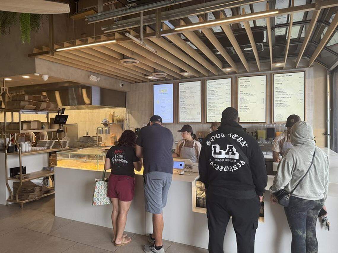A group of people standing at a counter and looking at a menu. The interior of the cafe has a modern, minimalist design with wooden accents and warm lighting. In the background, there are various food and drink menus on illuminated screens, along with display cases filled with pastries and other food items.