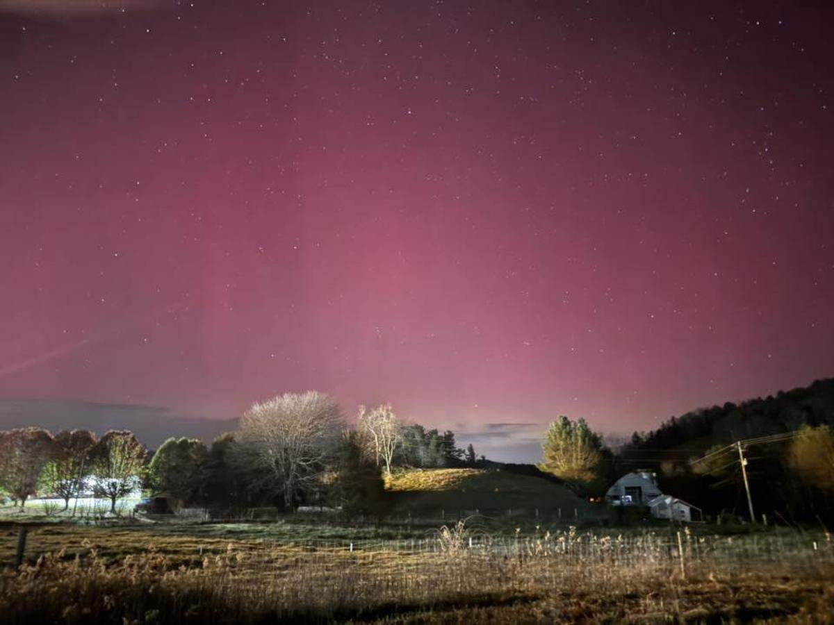 A wide, stunning nighttime landscape photo of a rural area under a deep magenta and pink sky, clearly visible with countless small stars. The aurora’s glow bathes the entire scene. In the middle ground, there are rolling hills, a few scattered trees, and a small white farmhouse nestled against a dark, tree-covered hillside. The immediate foreground is a field of dry, illuminated grasses.
