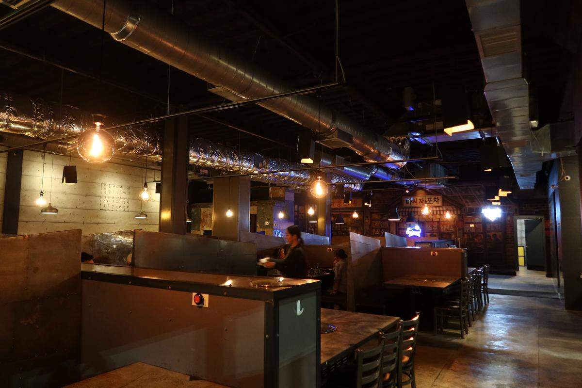 Another view of the dining area shows metal-partitioned booths and glowing globe lights that create a cozy, moody atmosphere. A staff member is seen walking through the spacious, concrete-floored restaurant near a mural of a Korean storefront.