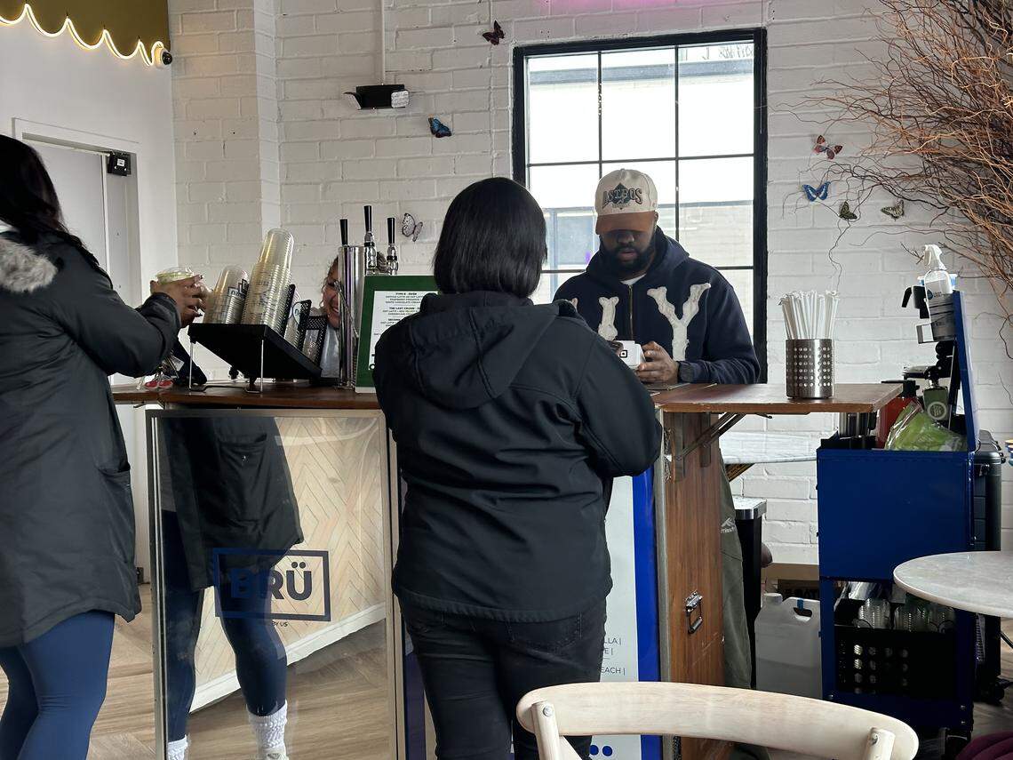 A barista looks down at a point-of-sale device while a customer in a black jacket stands at the counter. To the far left, another person’s hand is visible, holding up a green matcha drink. The scene is bright and airy with natural light coming from a large window.