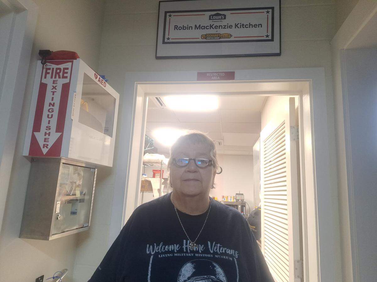 An individual with glasses stands in a doorway, looking directly at the camera, wearing a dark t-shirt that says “Welcome Home Veterans, Living Military History Museum.” Above the doorway, a sign reads “Lowe’s Robin Mackenzie Kitchen” with a logo underneath. To the left of the doorway, a red fire extinguisher and a white box for an AED are mounted on the wall. The room behind appears to be a kitchen.