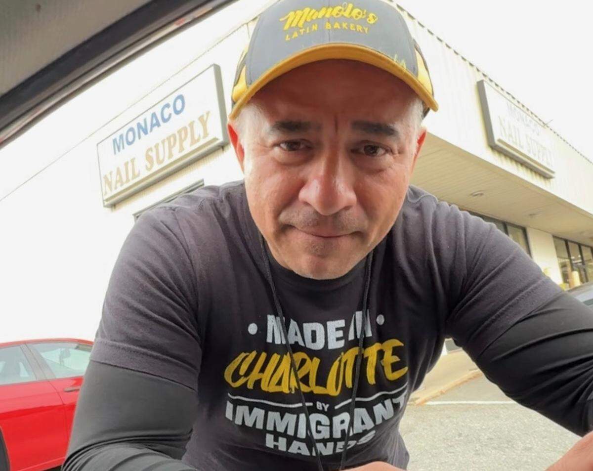 Close-up of a man speaking outdoors wearing a black t-shirt with ‘MADE IN CHARLOTTE BY IMMIGRANT HANDS’ in yellow text, and a baseball cap with ‘Manolo’s Latin Bakery’ visible on the front. A sign for ‘MONACO NAIL SUPPLY’ is visible in the background.