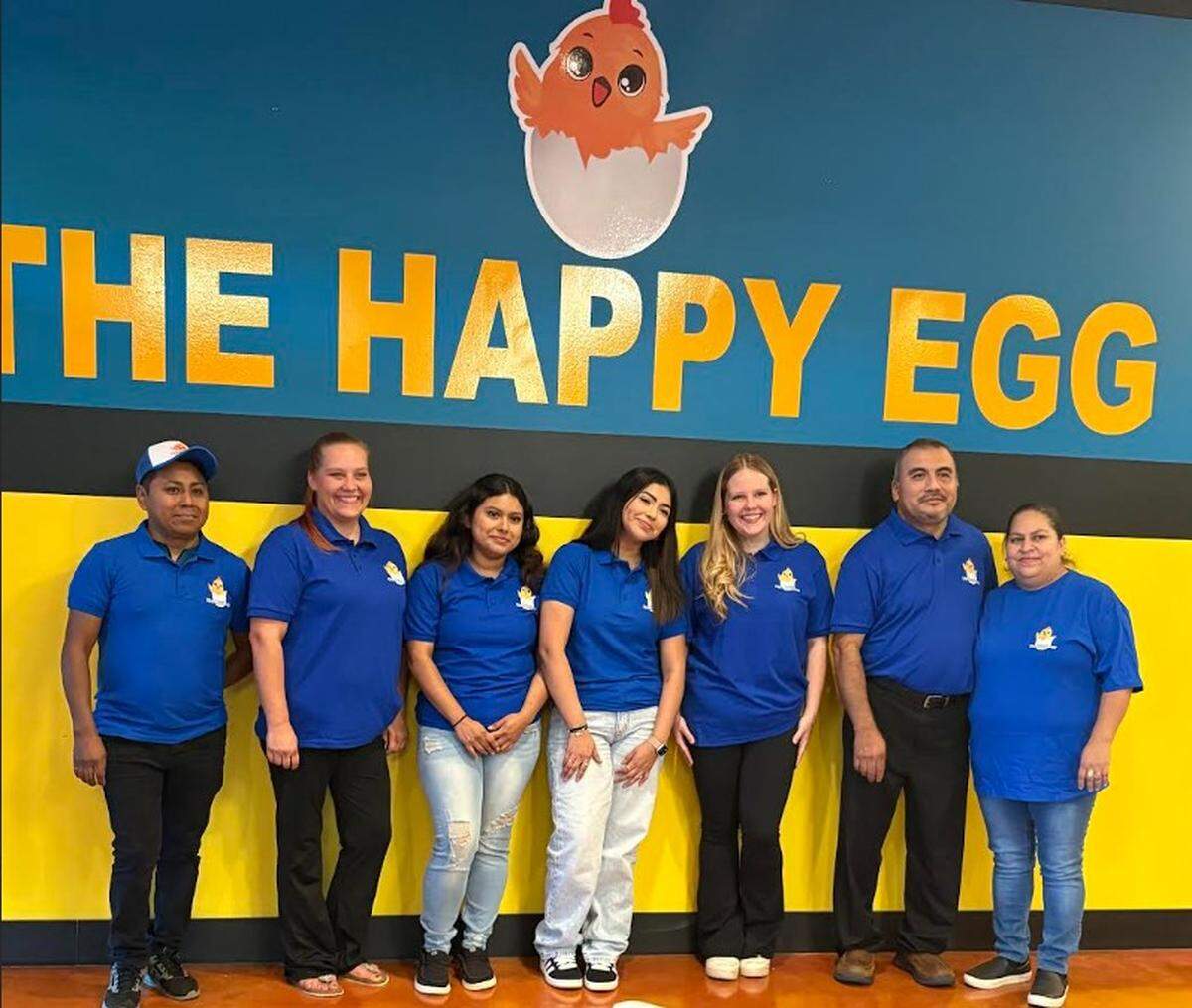 Seven staff members of “The Happy Egg” restaurant smiling and posing in front of a blue and yellow wall with the restaurant’s name and logo, which features a cartoon chick.