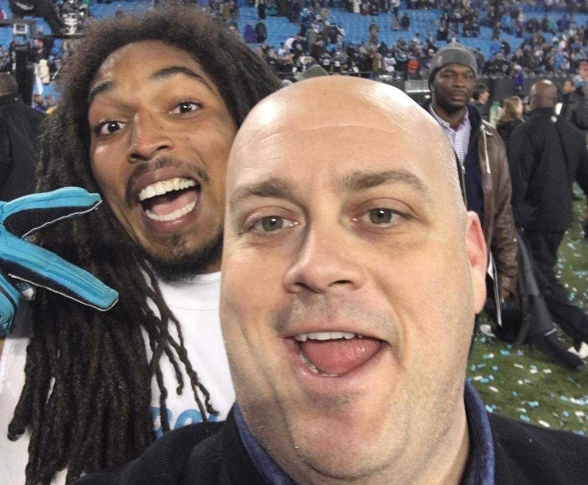 A man with a shaved head and a light beard is taking a selfie with a football player. The man is smiling and sticking out his tongue. Behind him, the football player, who has long dreadlocks and is wearing a white jersey and bright blue gloves, is also smiling broadly with his mouth open. The background is a blurred view of a crowded football stadium field and stands, with other people visible in the distance.