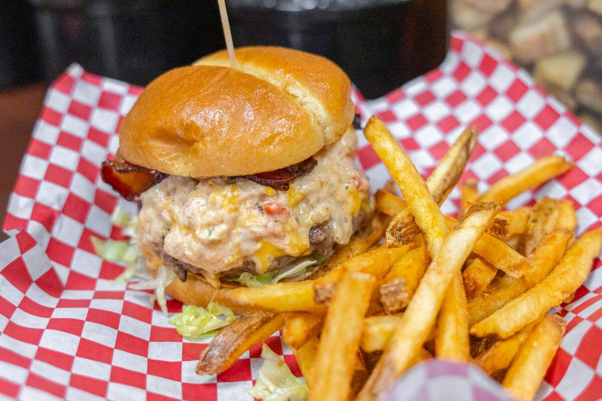 A close-up of a specialty burger topped with a generous scoop of pimento cheese and crispy bacon on a brioche bun. It is served in a checkered basket with a side of thick-cut, seasoned french fries.