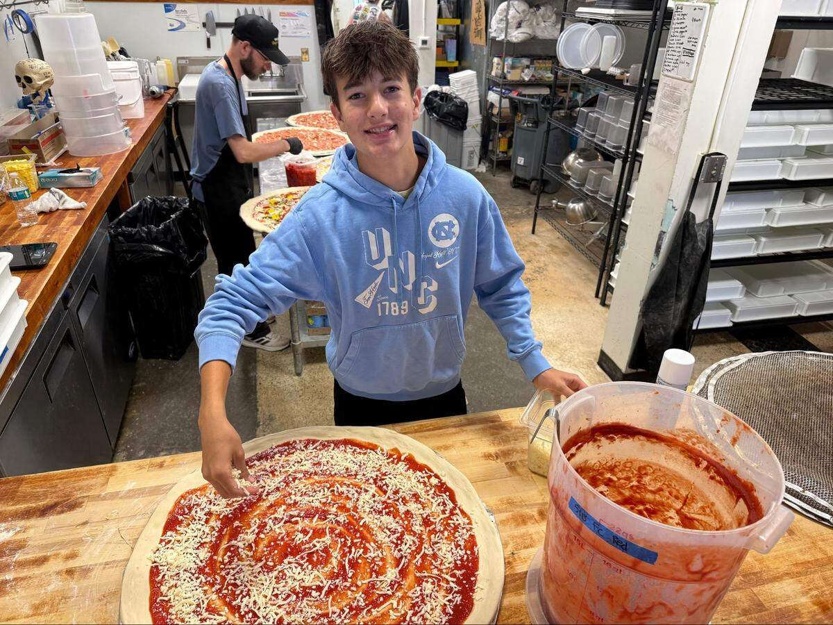 A high-angle, medium shot of a smiling teenager standing behind a wooden work counter in a commercial pizzeria kitchen, wearing a light blue UNC Chapel Hill hooded sweatshirt and is using a hand to sprinkle shredded mozzarella cheese over a massive, unbaked pizza crust spread with red sauce. To the right sits a large plastic bucket filled with pizza sauce. In the background, a kitchen worker wearing a black cap and apron prepares other large pizzas along a stainless steel counter.