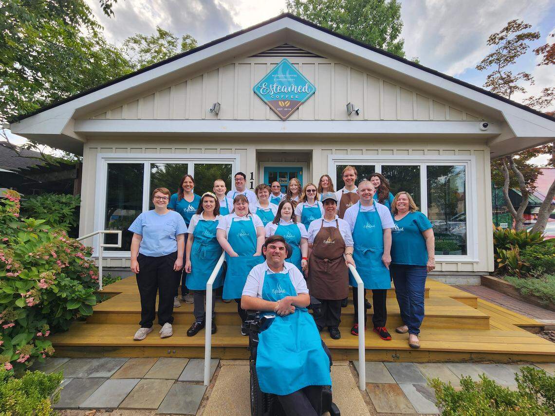 A large group of employees stands together on the wooden porch in front of Esteamed Coffee, a light-colored building with a teal logo. Most of the staff are wearing bright teal aprons or shirts, smiling for a group portrait outdoors.