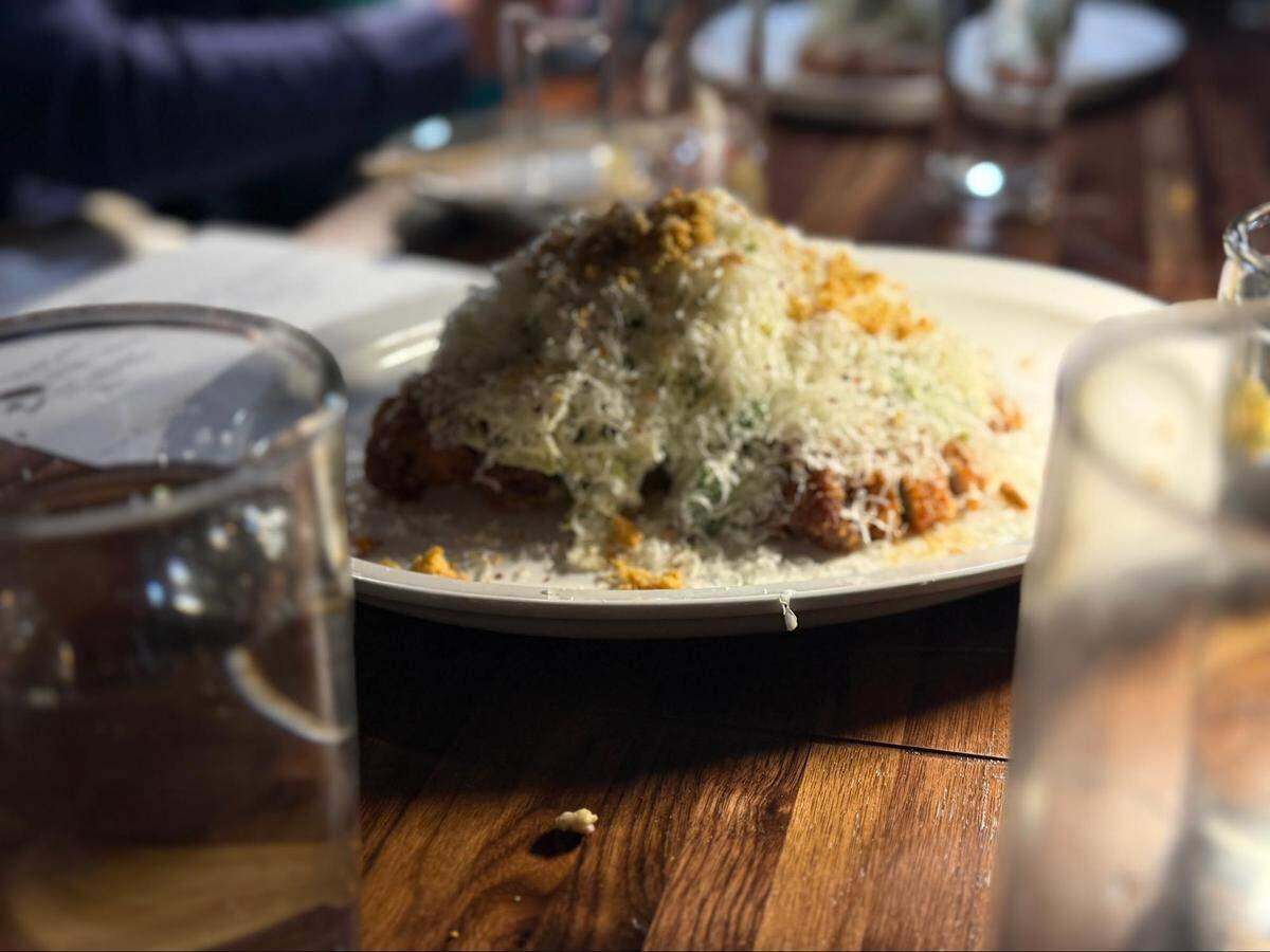 A side view of a crispy fried chicken cutlet or katsu, almost entirely obscured by a tall, fluffy mound of finely shredded white cheese. The dish is served on a white plate on a dark wood table.